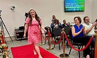  Woman in pink dress walks down a red carpet while people are clapping in the audience.
